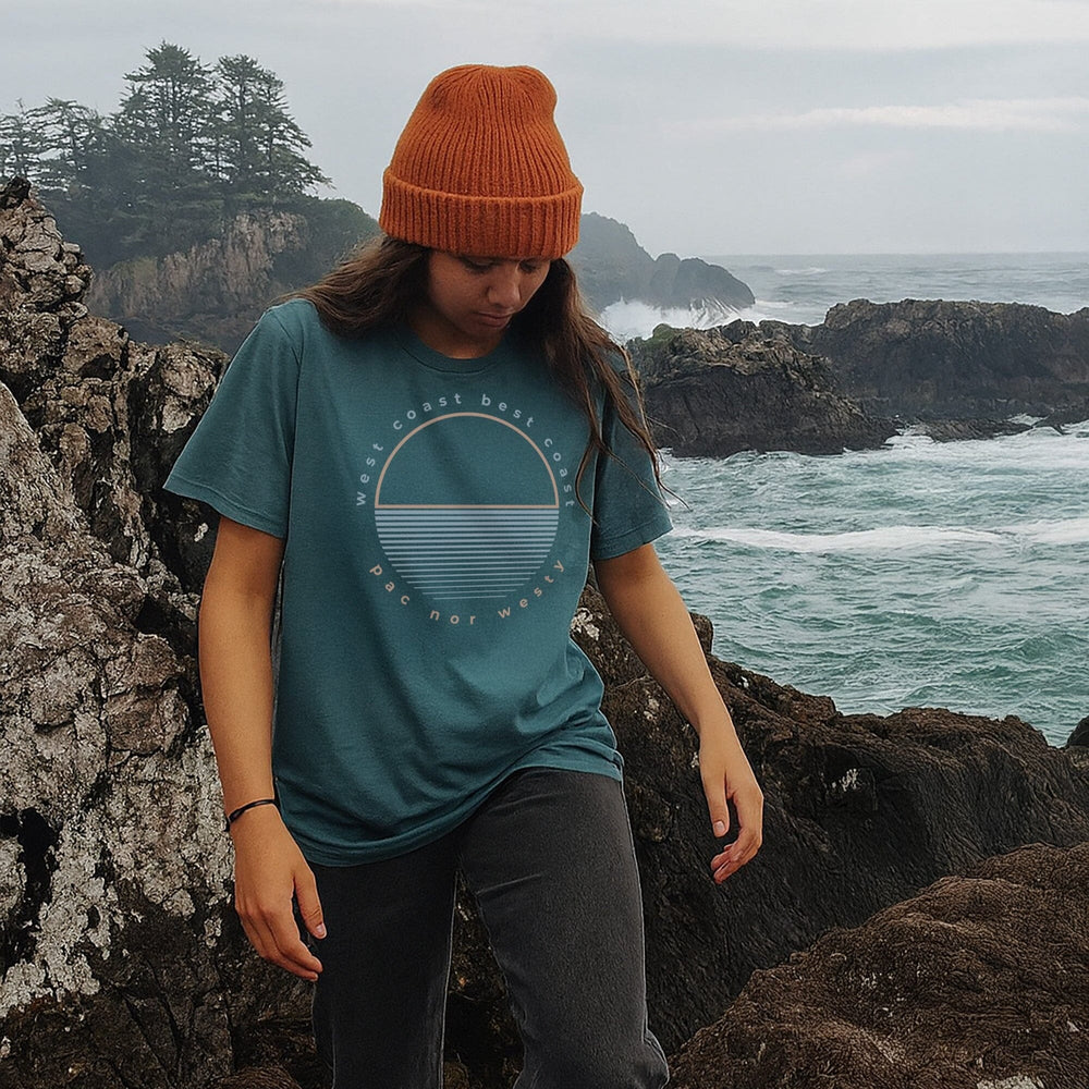 Person wearing a teal t-shirt with a logo, orange beanie, and black sneakers standing on rocky coastal terrain.